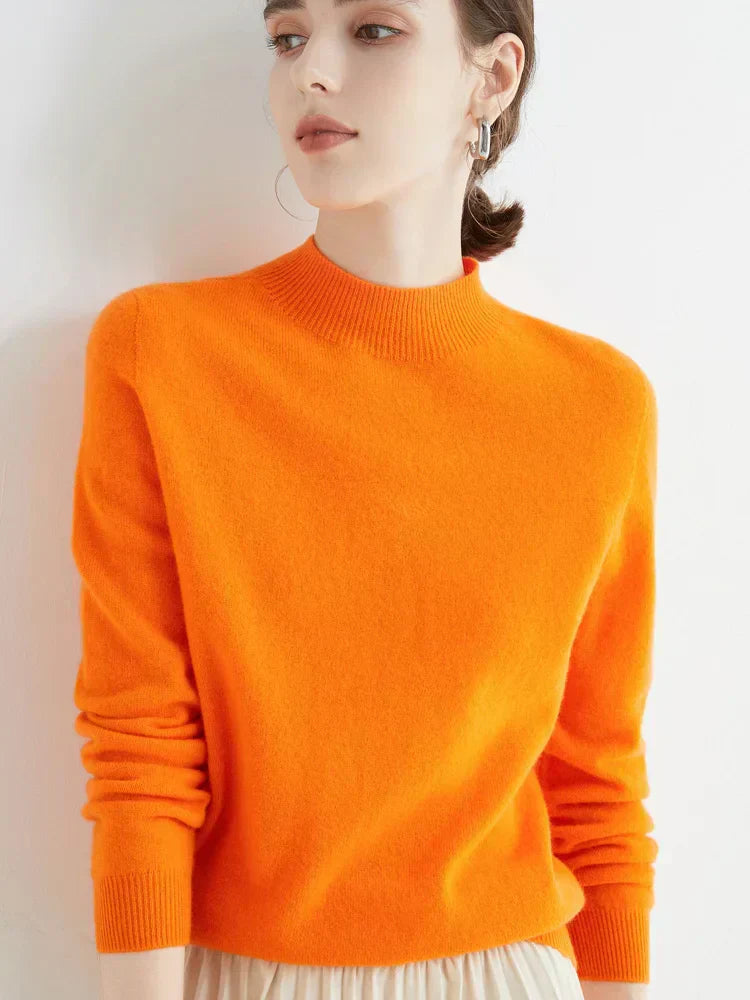Femme portant un pull orange vif en cachemire, manches longues, col rond, posant contre un mur blanc. Mode automne-hiver, style élégant et moderne.