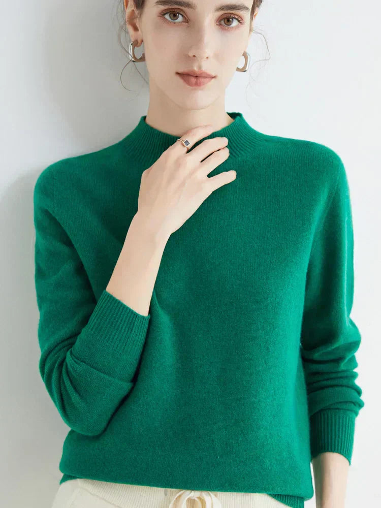 Femme portant un pull vert en cachemire, manches longues, col rond, posant contre un fond blanc. Mode automne-hiver, élégance et confort.