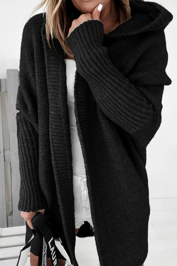 Femme portant un cardigan noir en tricot épais, manches longues, capuche, tenue décontractée, intérieur moderne, style automne-hiver tendance.