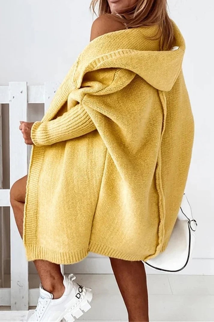 Femme portant un cardigan jaune oversize en tricot, assise de dos, avec des baskets blanches et un sac à main blanc. Mode automne-hiver tendance.