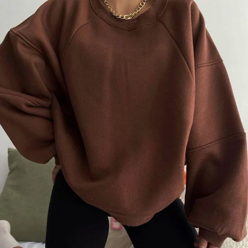 Femme portant un pull marron oversize, manches longues, avec un collier doré, posant sur un canapé vert. Mode décontractée automne-hiver.