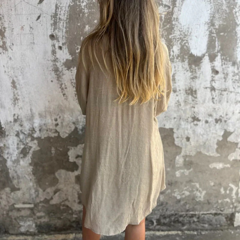 Femme aux cheveux blonds portant une robe beige en lin, debout devant un mur texturé gris. Mode décontractée, style minimaliste, arrière-plan urbain.