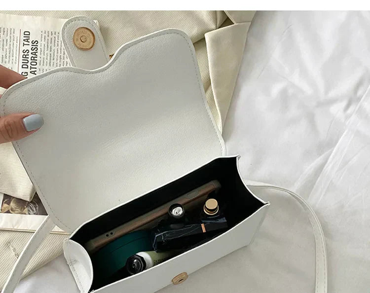 Sac à main blanc ouvert avec rouge à lèvres, parfum et stylo à l'intérieur, posé sur un lit avec un magazine. Accessoire mode élégant et pratique.