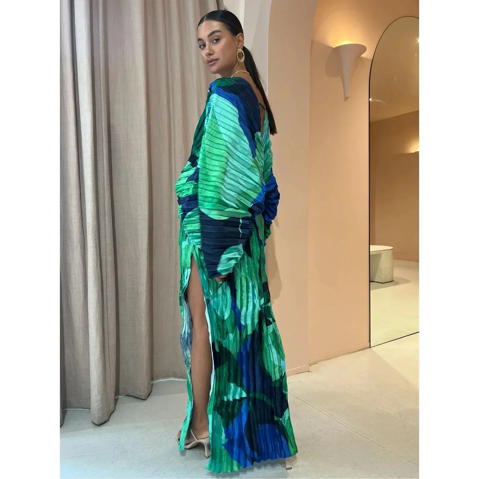 Femme portant une robe longue plissée verte et bleue avec motifs abstraits, posant dans un intérieur moderne. Mode élégante, tendance 2023.