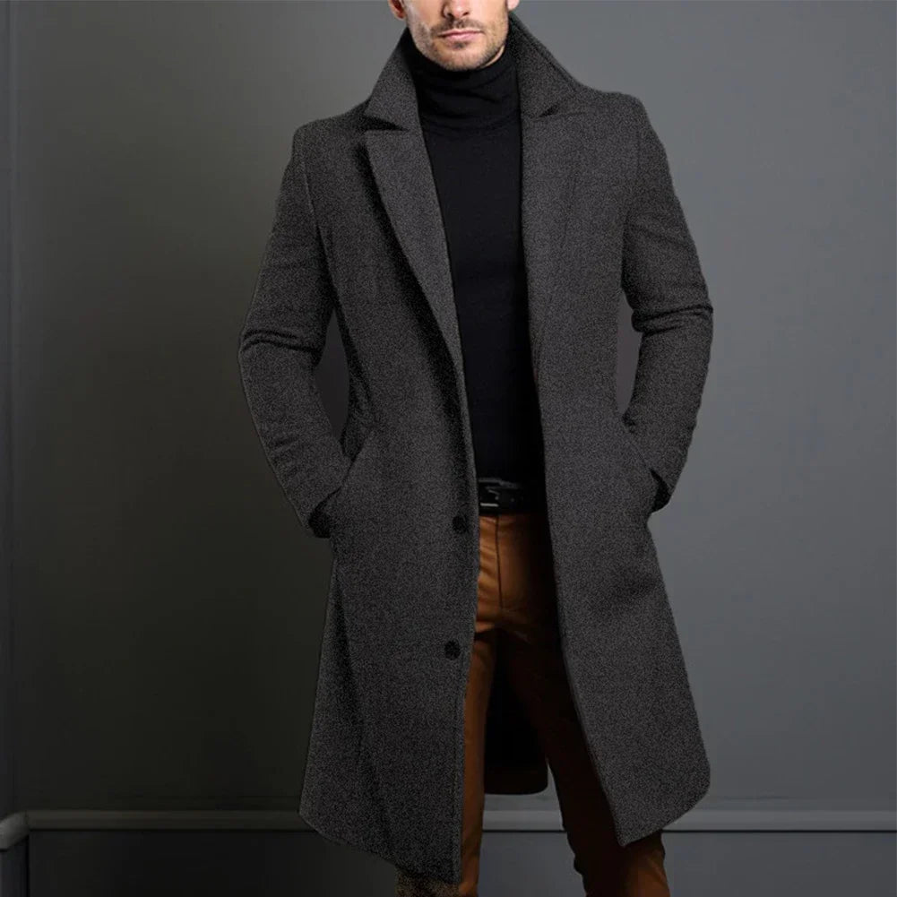 Homme portant un manteau gris élégant, col relevé, sur un pull noir et un pantalon marron, posant contre un mur gris. Mode masculine chic.
