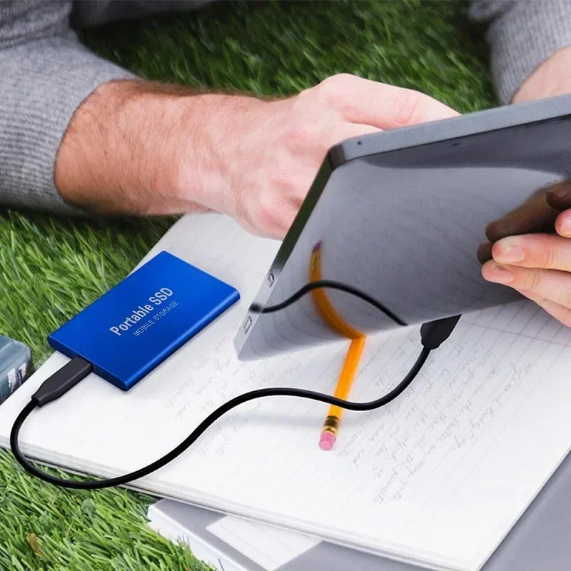 Homme utilisant une tablette connectée à un SSD portable bleu sur un cahier, sur fond de gazon. Stockage externe, technologie mobile, travail nomade.