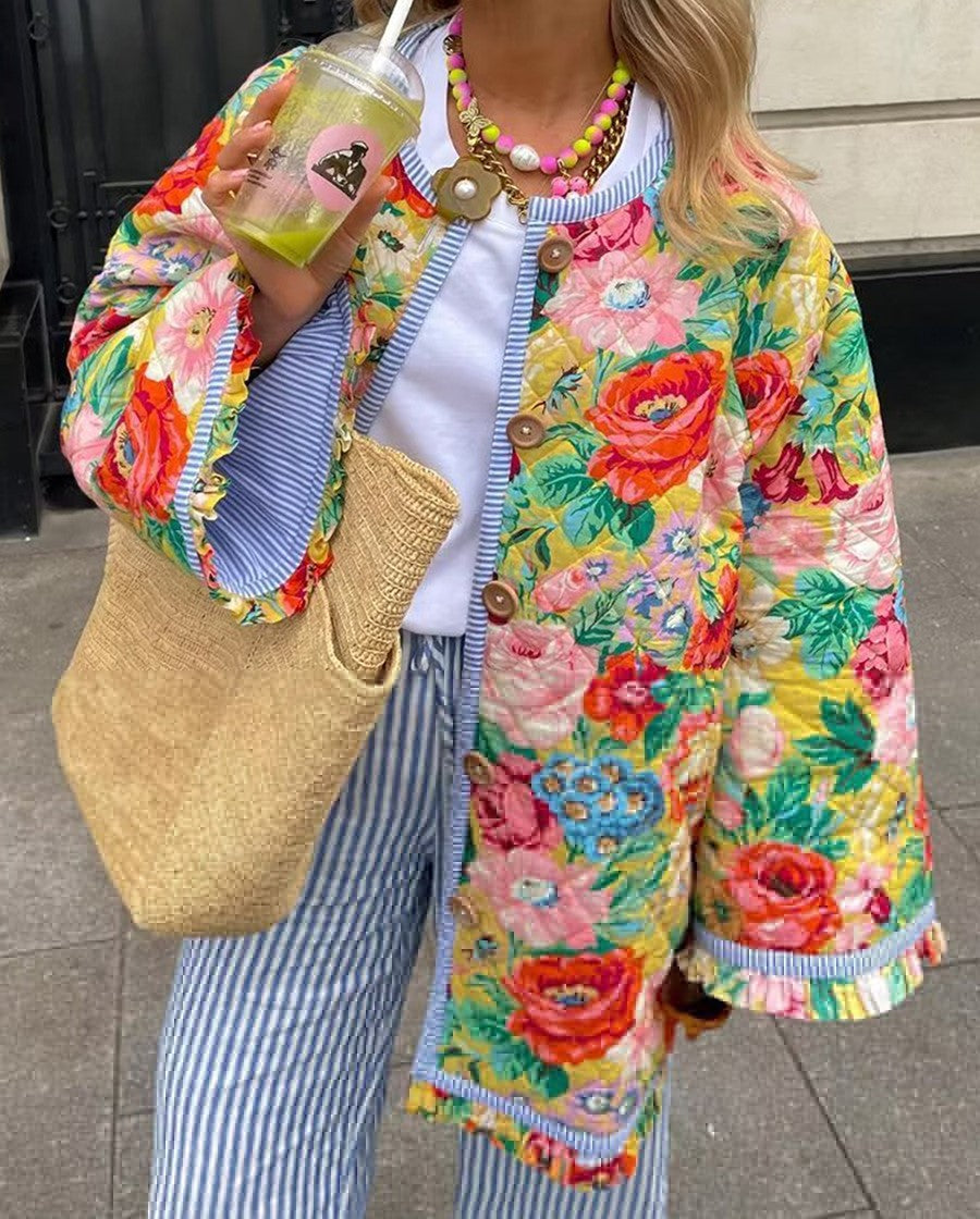 Femme portant un manteau fleuri coloré, tenant un sac en paille et une boisson. Mode printemps, style bohème, rue urbaine.