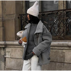 Femme en manteau gris et bonnet blanc tenant un croissant, debout devant un bâtiment ancien. Mode hivernale, style urbain, Paris.