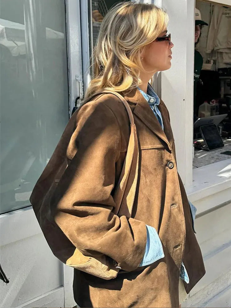 Femme blonde portant un manteau en daim marron et lunettes de soleil, debout à l'extérieur d'un café. Mode automne, style décontracté chic.