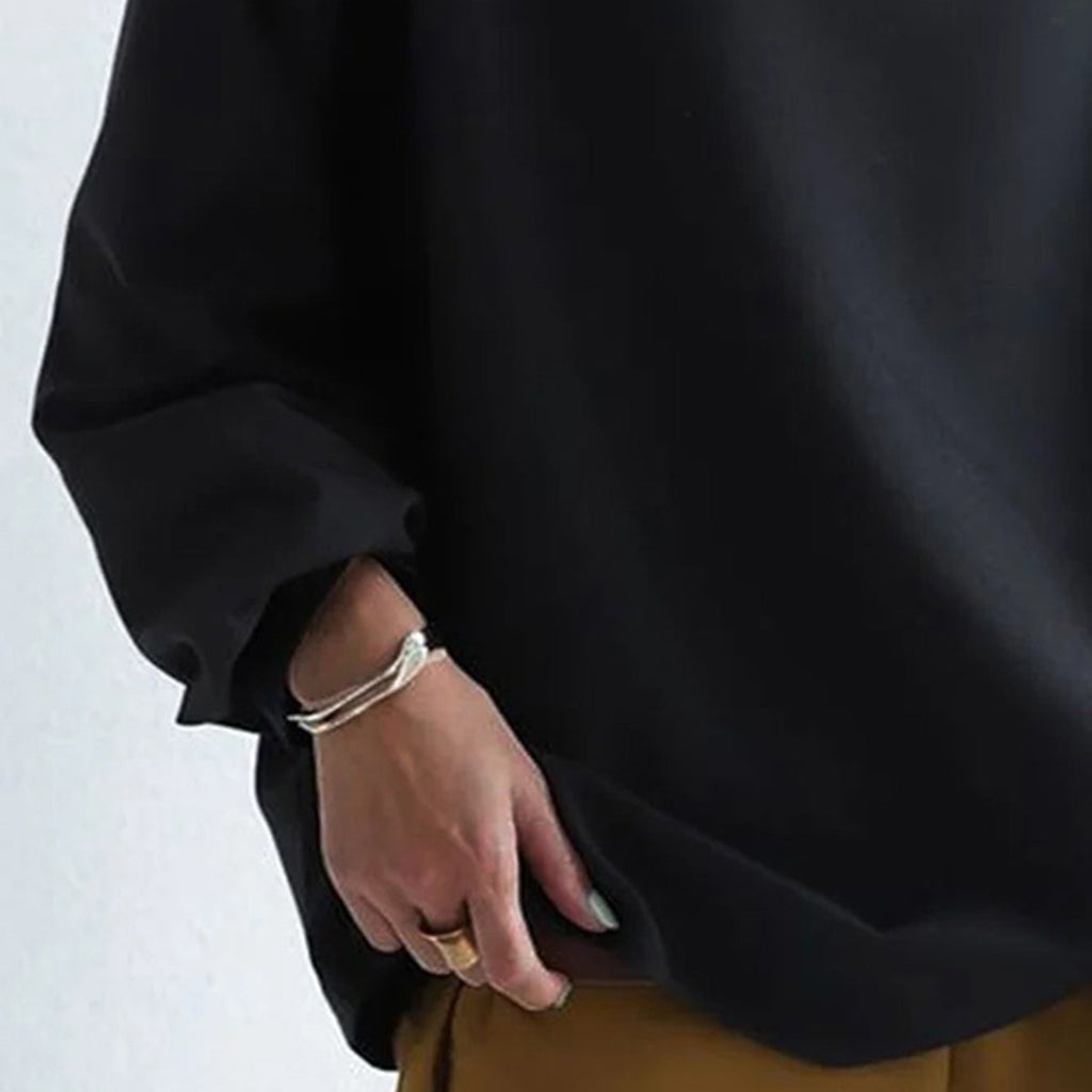 Personne portant un pull noir ample, tenant l'ourlet avec une main ornée de bagues et d'un bracelet, pantalon marron visible. Mode décontractée élégante.