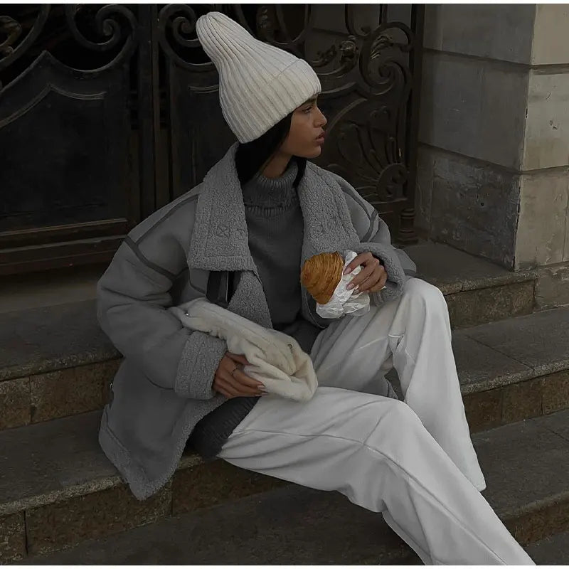 Femme assise sur des marches, portant un manteau gris et un bonnet blanc, tenant un croissant. Mode hivernale, style urbain, vêtements chauds.