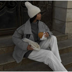 Femme assise sur des marches, portant un manteau gris et un bonnet blanc, tenant un croissant. Mode hivernale, style urbain, vêtements chauds.