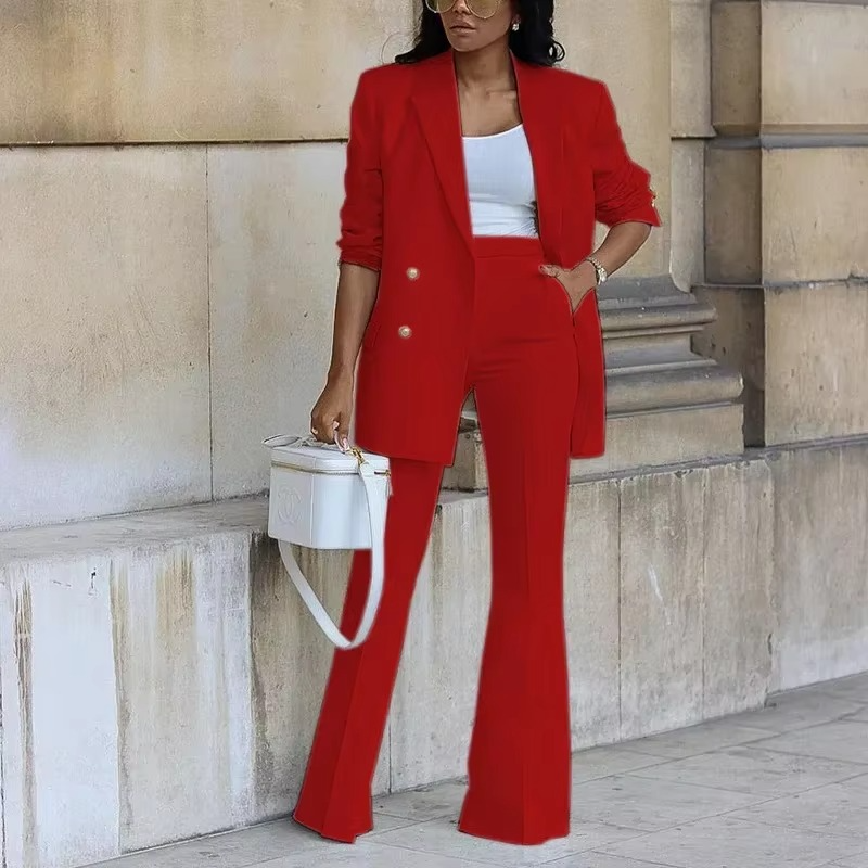 Femme en tailleur rouge élégant, veste à boutons dorés, pantalon large, top blanc, sac blanc, pose devant un mur en pierre. Mode chic et moderne.