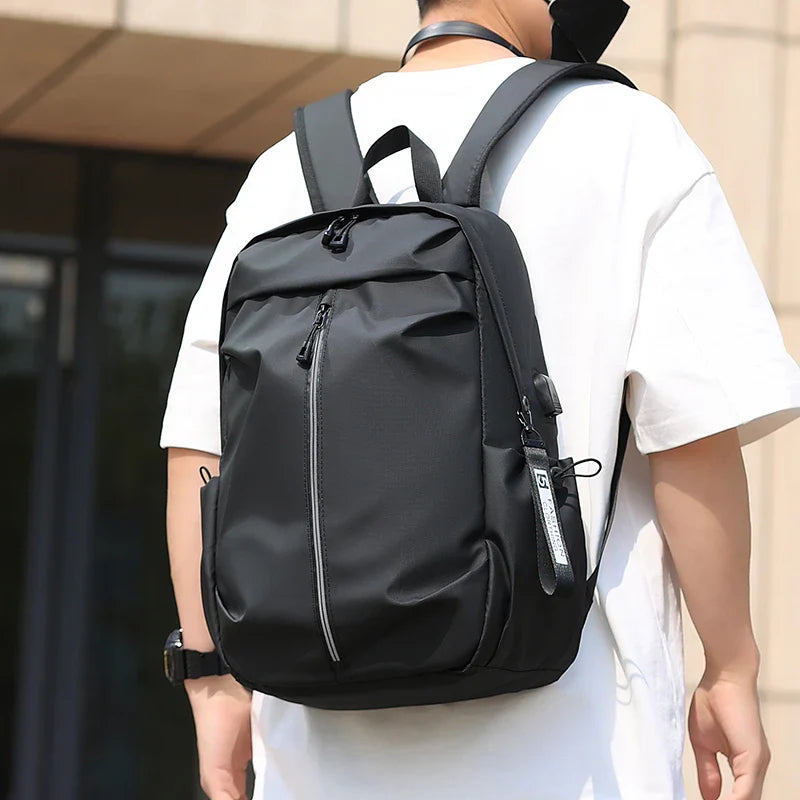 Sac à dos noir moderne porté par une personne en t-shirt blanc, design urbain, idéal pour le voyage et l'école, avec fermeture éclair et poches latérales.