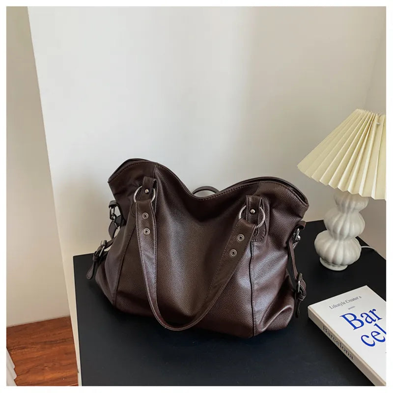 Sac à main en cuir marron sur une table noire, à côté d'une lampe blanche et d'un livre intitulé "Barcelona". Accessoire mode élégant et pratique.