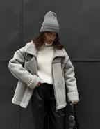Femme portant un manteau gris en laine, pull blanc, bonnet gris et lunettes de soleil, posant devant un mur noir. Mode hiver, style urbain.