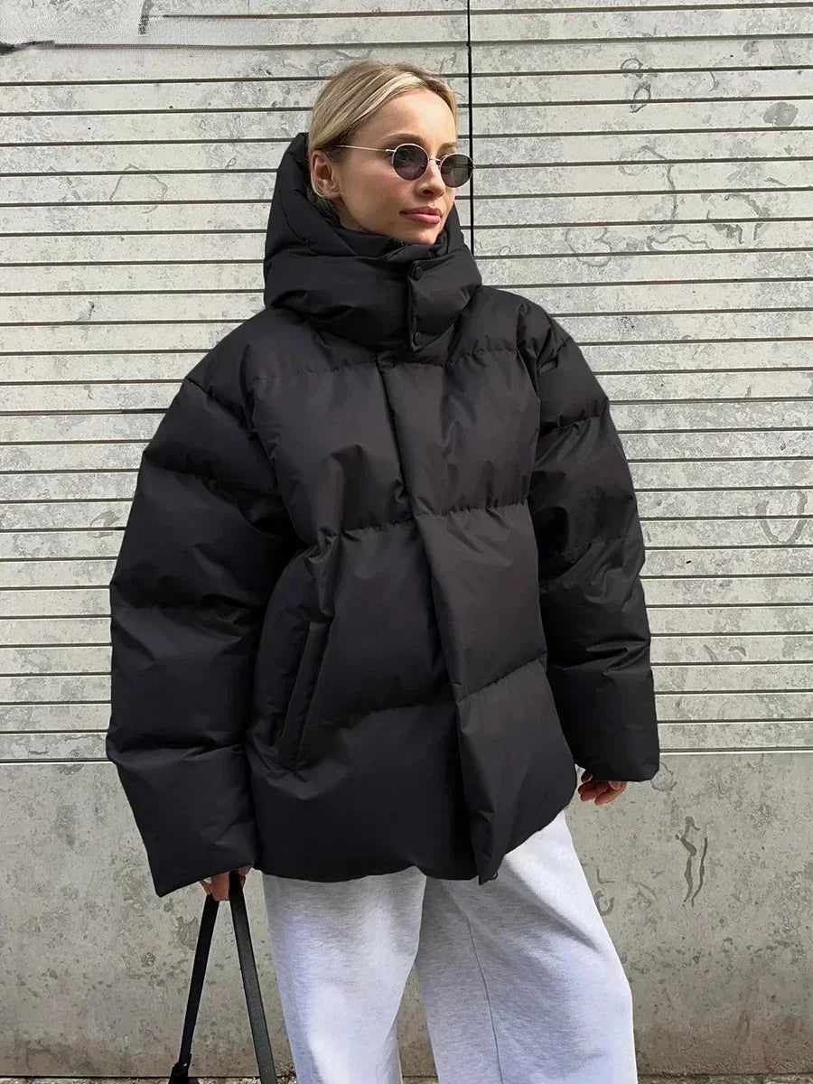 Femme portant une doudoune noire oversize, lunettes de soleil rondes, pantalon gris, devant un mur rayé. Mode hiver, style urbain tendance.