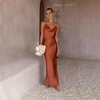 Robe de soirée longue en satin marron, femme souriante tenant un bouquet de fleurs blanches, fond minimaliste en pierre beige.