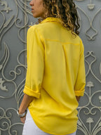 Femme de dos portant une chemise jaune vif, manches retroussées, devant un mur décoratif gris. Mode féminine, style décontracté, élégance moderne.