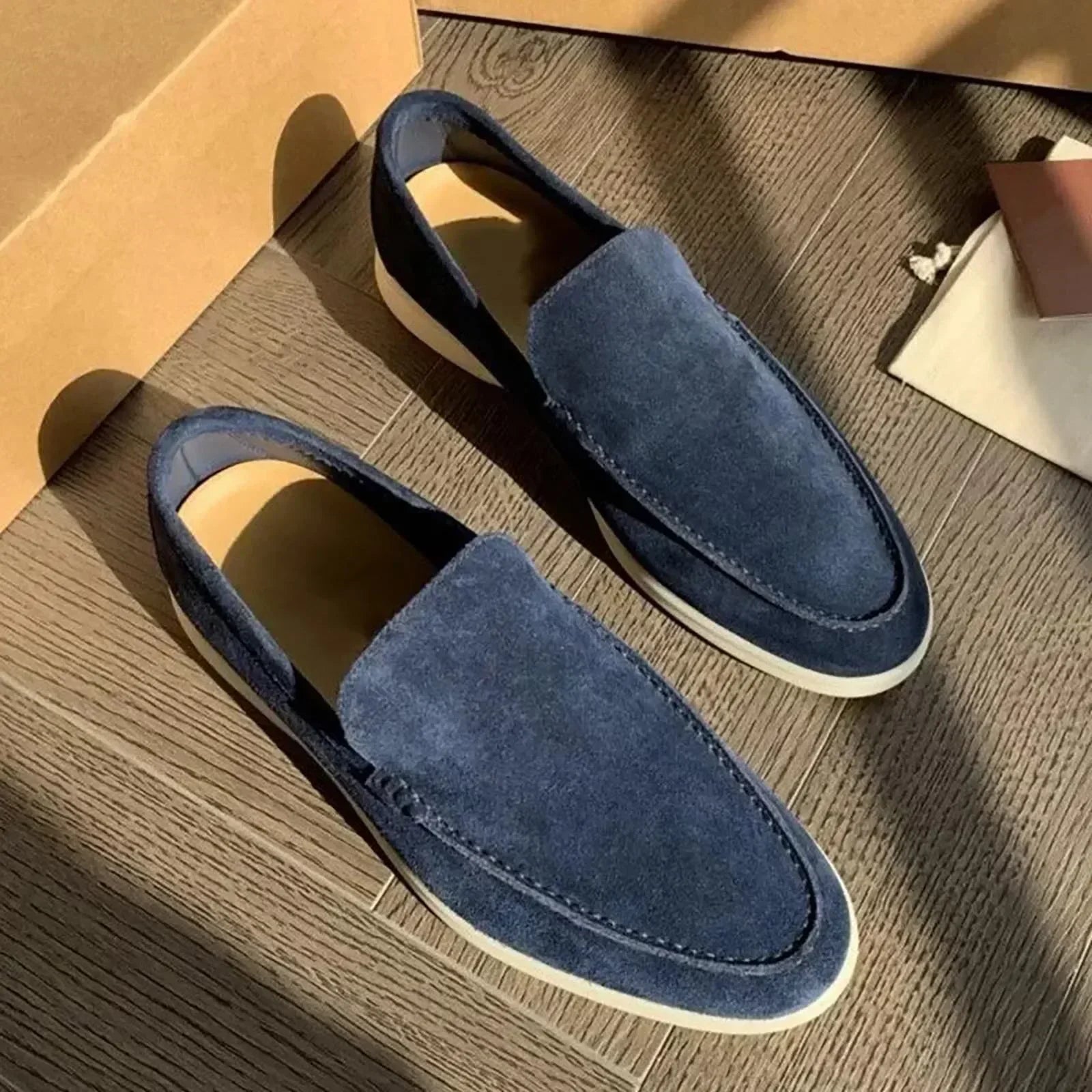 Mocassins en daim bleu pour homme sur parquet en bois, éclairés par la lumière du soleil. Chaussures élégantes et confortables, style décontracté.