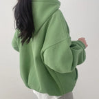 Femme portant un sweat à capuche vert ample, style décontracté, mode automne-hiver, dos tourné, cheveux longs noirs, intérieur minimaliste.