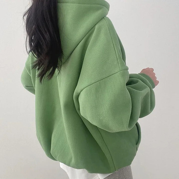 Femme portant un sweat à capuche vert ample, style décontracté, mode automne-hiver, dos tourné, cheveux longs noirs, intérieur minimaliste.