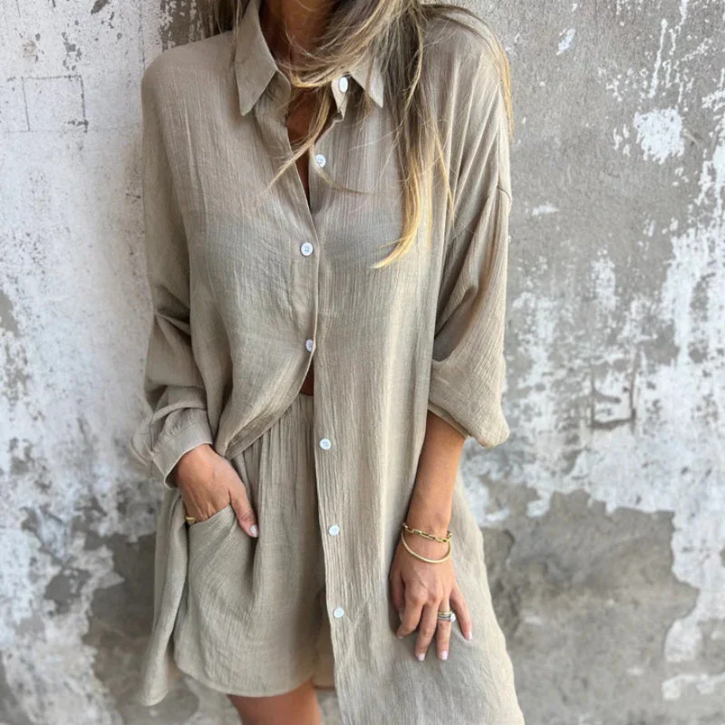 Femme portant une chemise longue beige en lin, manches retroussées, devant un mur texturé. Mode décontractée, style estival chic.