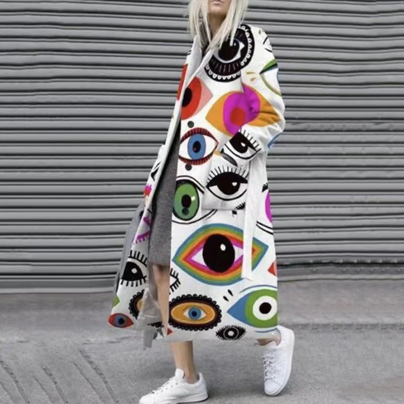 Femme portant un manteau long blanc avec motifs d'yeux colorés, debout devant un mur gris ondulé, chaussures blanches, style moderne et audacieux.