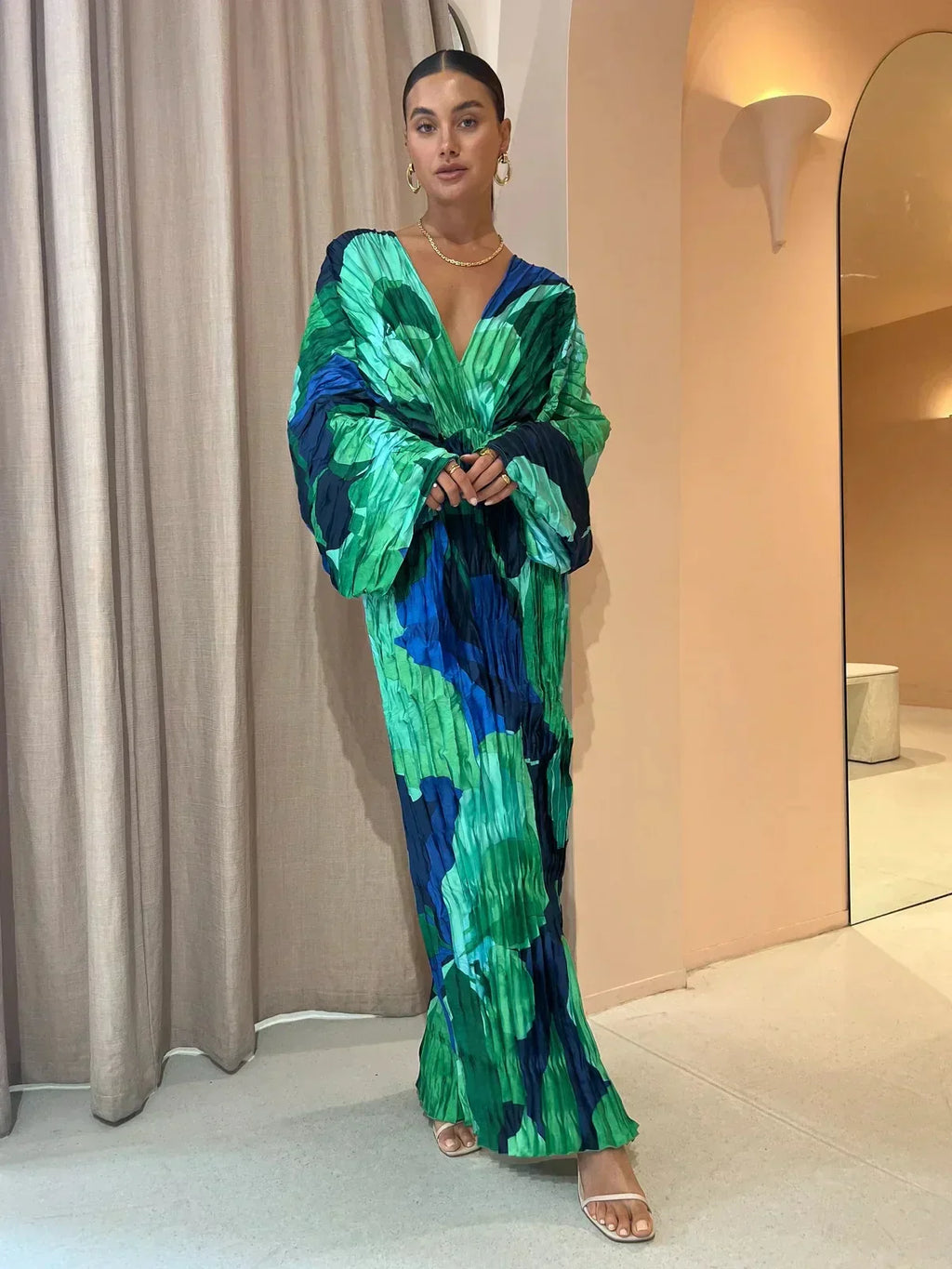 Robe longue plissée verte et bleue, manches larges, portée par une femme élégante dans un intérieur moderne. Mode féminine, style chic et tendance.