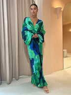 Robe longue plissée verte et bleue, manches larges, portée par une femme élégante dans un intérieur moderne. Mode féminine, style chic et tendance.