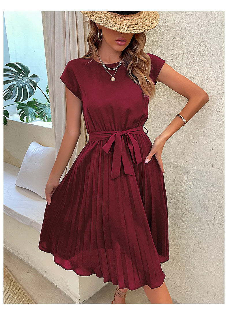 Robe bordeaux plissée élégante portée par une femme avec chapeau de paille, collier et bracelet, posant dans un intérieur lumineux avec plante verte.
