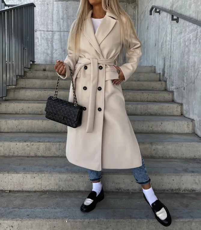Femme en trench-coat beige, jeans, mocassins noirs et chaussettes blanches, tenant un sac à main noir matelassé, posant sur des escaliers en béton.