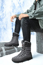 Bottes d'hiver grises pour homme avec doublure en fourrure, portées avec un jean noir et une veste verte, posées sur un fond enneigé.