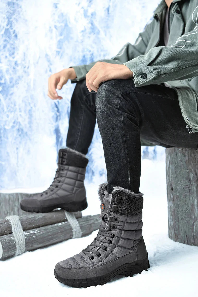 Bottes d'hiver grises pour homme avec doublure en fourrure, portées avec un jean noir et une veste verte, posées sur un fond enneigé.