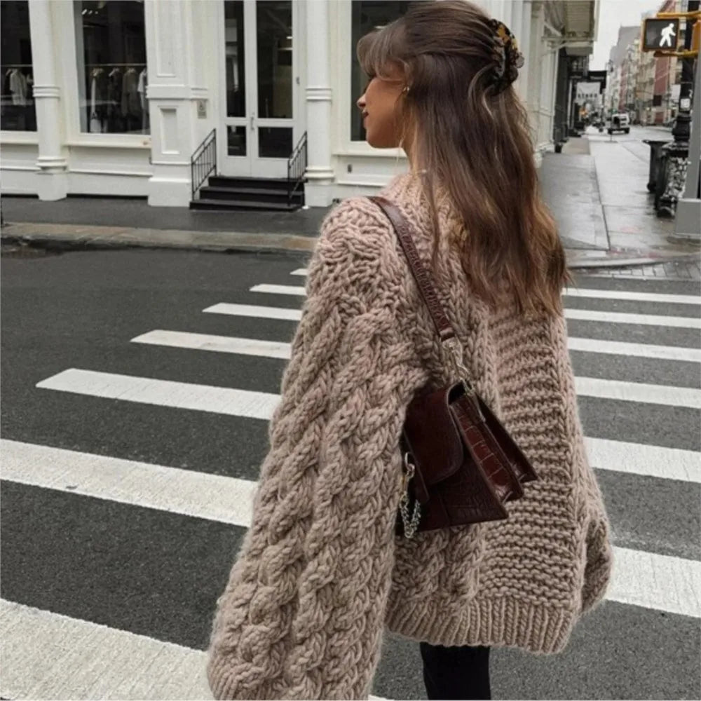 Jeune femme portant un pull en maille épaisse beige et un sac à main marron, traversant une rue urbaine. Mode automne, style décontracté chic.