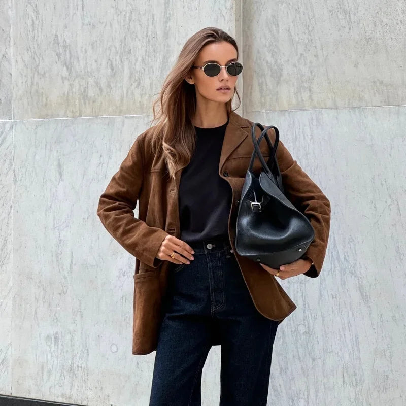 Femme élégante portant un manteau marron, lunettes de soleil et sac à main noir, posant devant un mur en marbre. Mode automne chic et tendance.