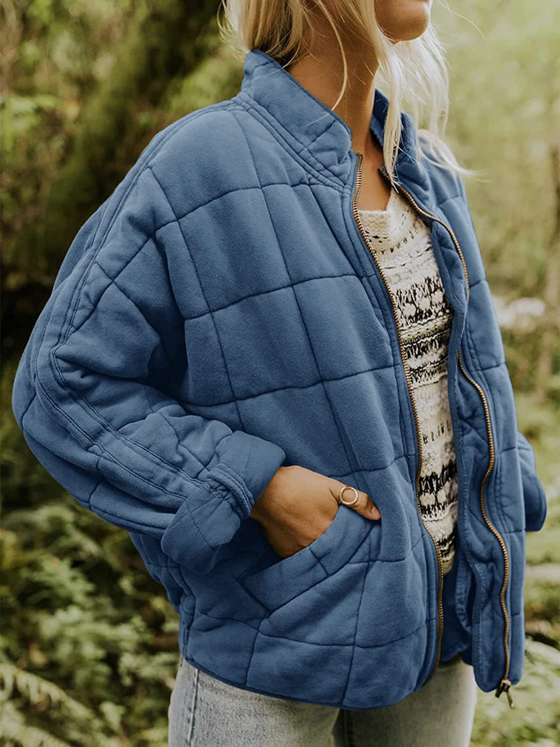 Femme portant une veste matelassée bleue, manches longues, sur un pull à motifs, dans un cadre naturel. Mode automne, style décontracté.