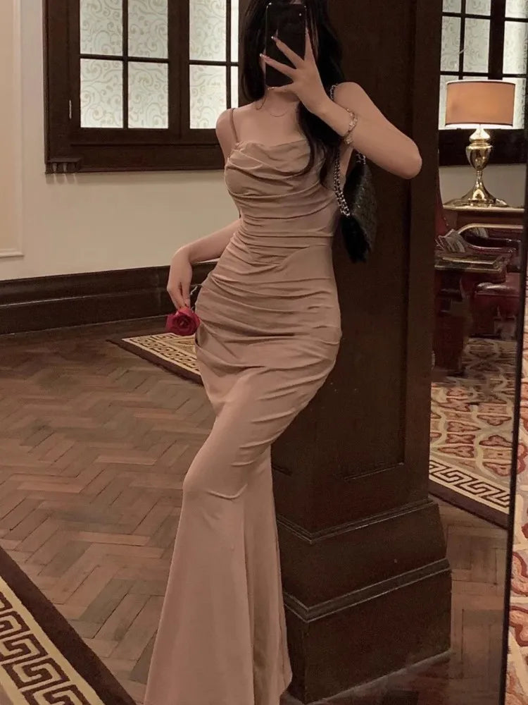 Femme en robe longue beige élégante, tenant une rose rouge, posant dans un intérieur chic avec parquet et lampe allumée. Mode soirée glamour.