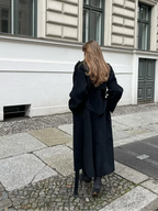 Femme portant un long manteau noir, marchant sur un trottoir pavé devant un bâtiment historique. Mode automne, style urbain élégant.
