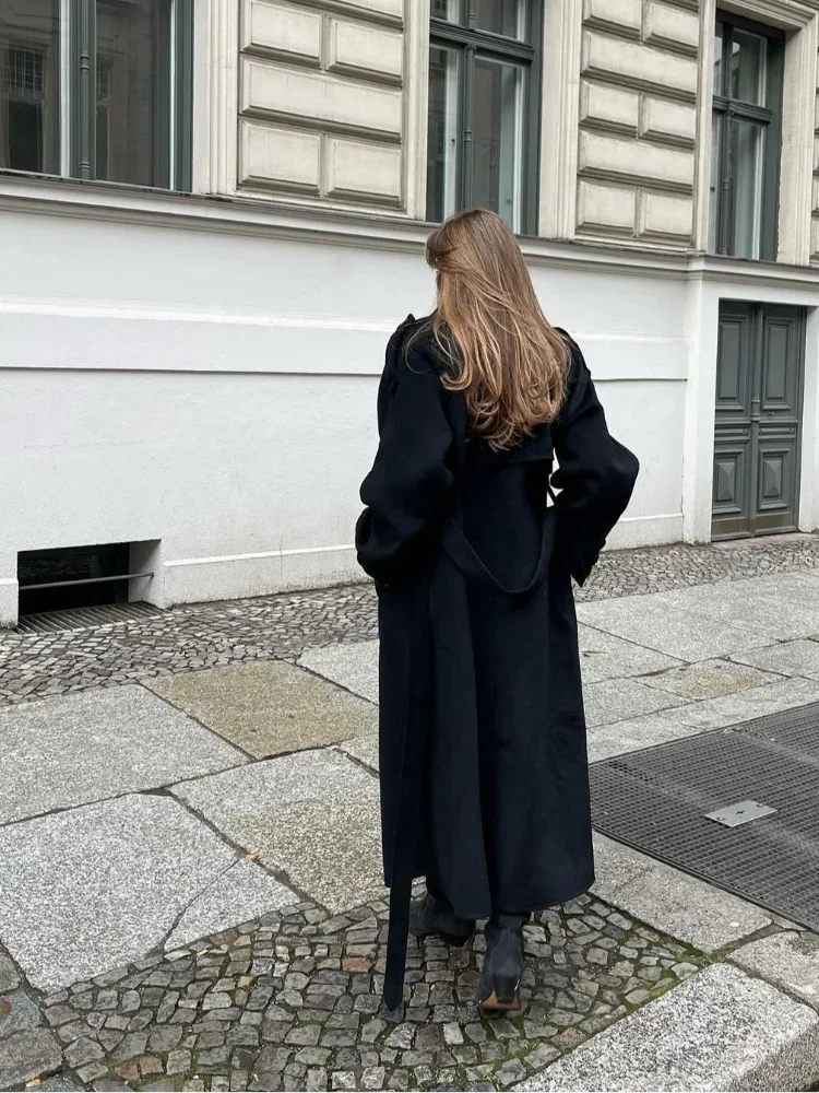Femme portant un long manteau noir, marchant sur un trottoir pavé devant un bâtiment historique. Mode automne, style urbain élégant.
