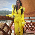 Femme en combinaison de ski jaune vif avec fourrure, posant sur un balcon en bois, paysage enneigé en arrière-plan. Mode hiver, tenue de ski élégante.