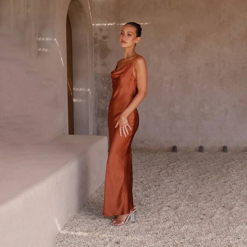 Femme en robe longue satinée marron, posant dans un intérieur minimaliste avec murs texturés et sol en gravier, style élégant et moderne.