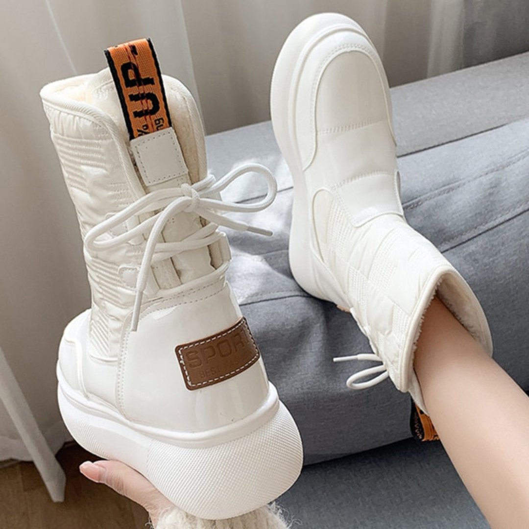 Bottes blanches en cuir synthétique avec semelle épaisse, lacets et étiquette marron "SPORT", posées sur un canapé gris. Mode tendance hiver.