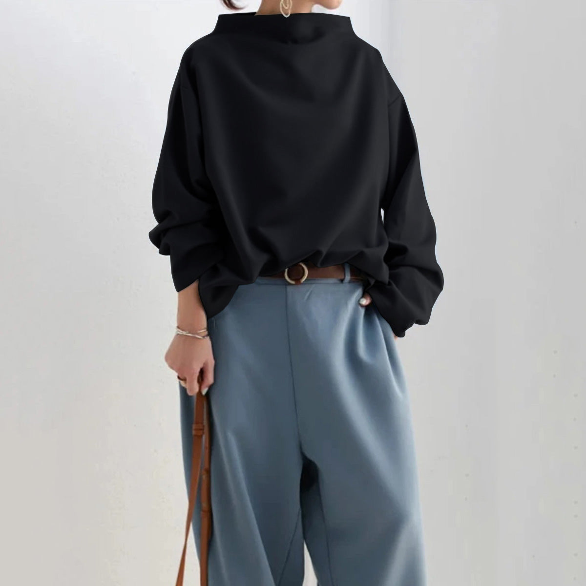 Personne portant un pull noir ample et un pantalon bleu taille haute, accessoirisé d'une ceinture marron et d'un sac à main. Mode décontractée élégante.
