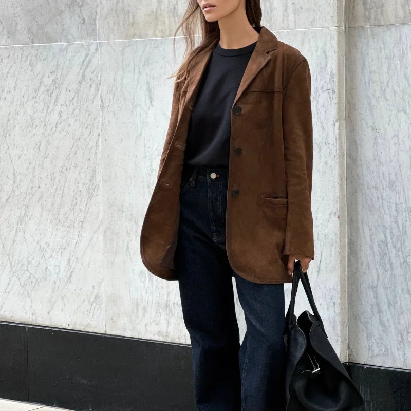 Femme portant un blazer marron en daim, t-shirt noir et jean, tenant un sac noir, posant devant un mur en marbre. Mode automne chic et élégante.