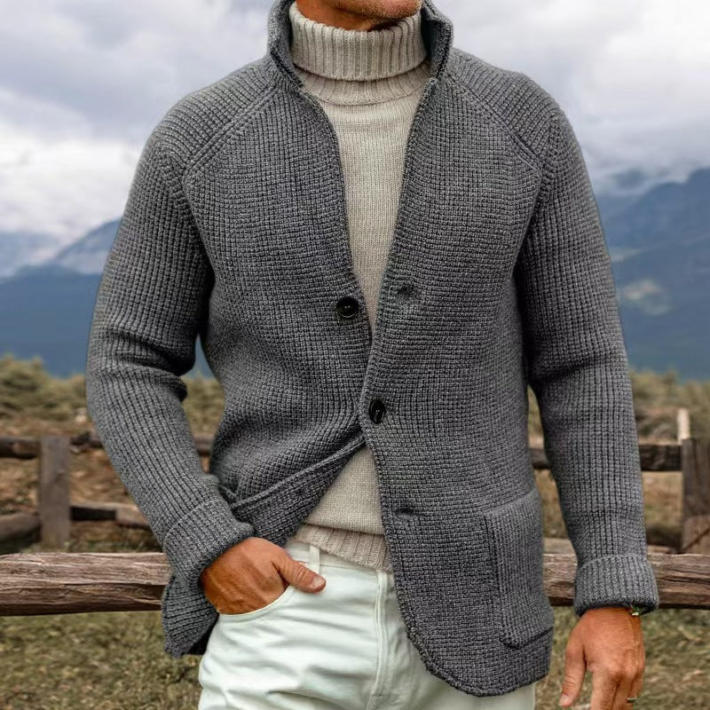 Homme portant un cardigan gris en laine avec col montant, sur un pull beige, debout devant un paysage montagneux. Mode masculine automne-hiver.