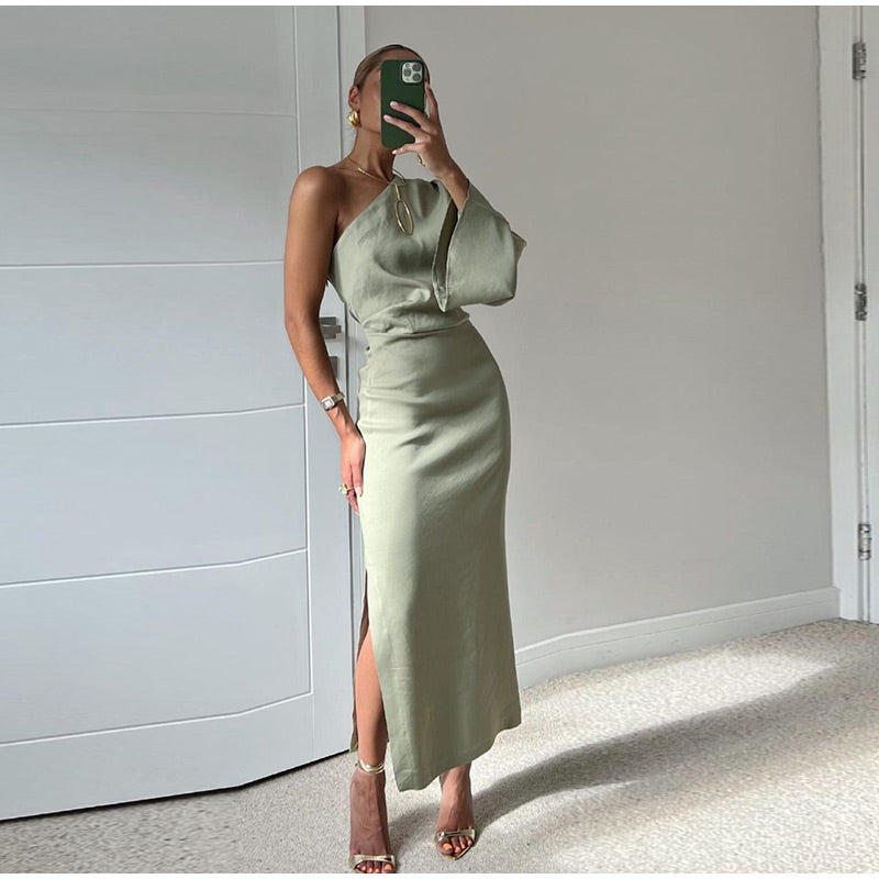 Robe longue verte asymétrique avec fente, femme tenant un smartphone, pose élégante, intérieur moderne, mode tendance, style chic.