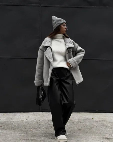 Femme portant un manteau gris en laine, pull blanc, pantalon noir ample et bonnet gris, posant devant un mur noir. Mode hiver chic et moderne.