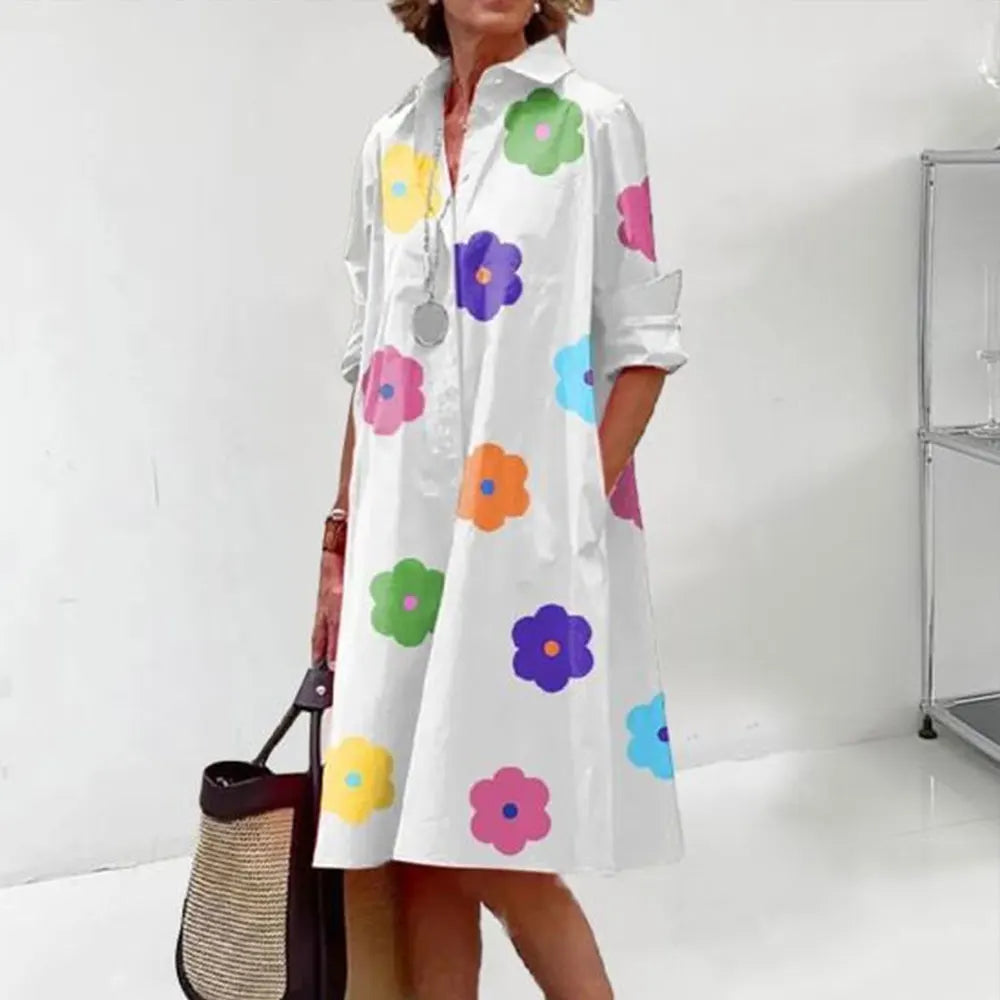 Robe chemise blanche à motifs floraux colorés, manches longues retroussées, portée par une femme tenant un sac en osier. Mode féminine tendance.