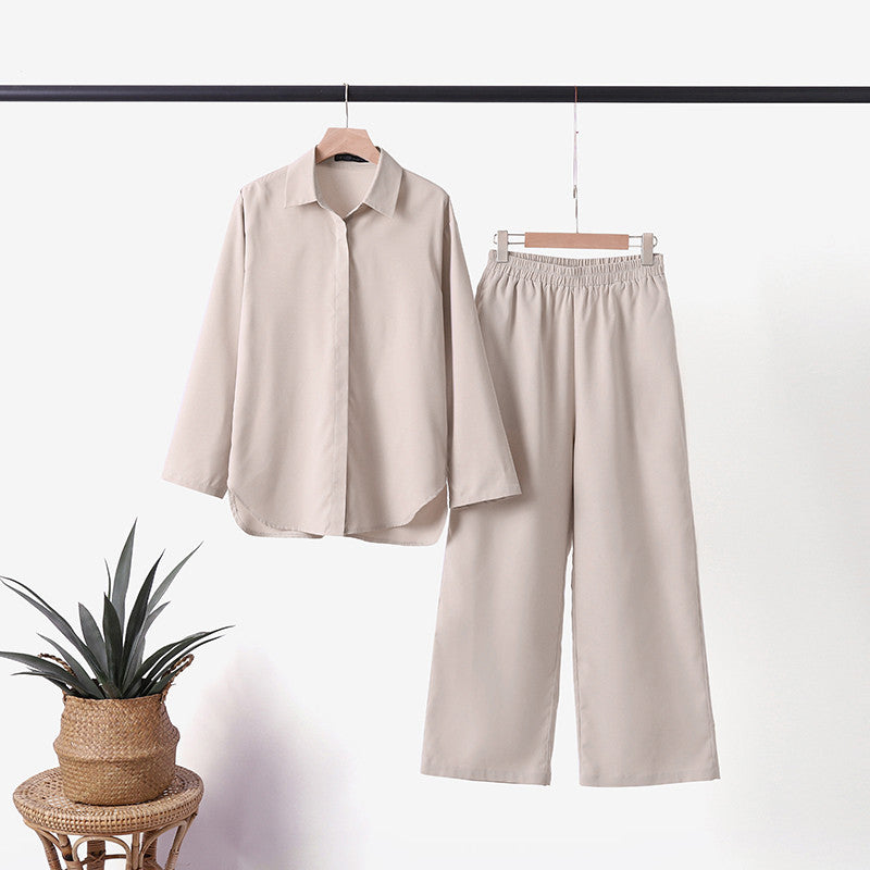 Tenue élégante beige composée d'une chemise à manches longues et d'un pantalon ample, suspendue sur un cintre, avec une plante en pot à côté.